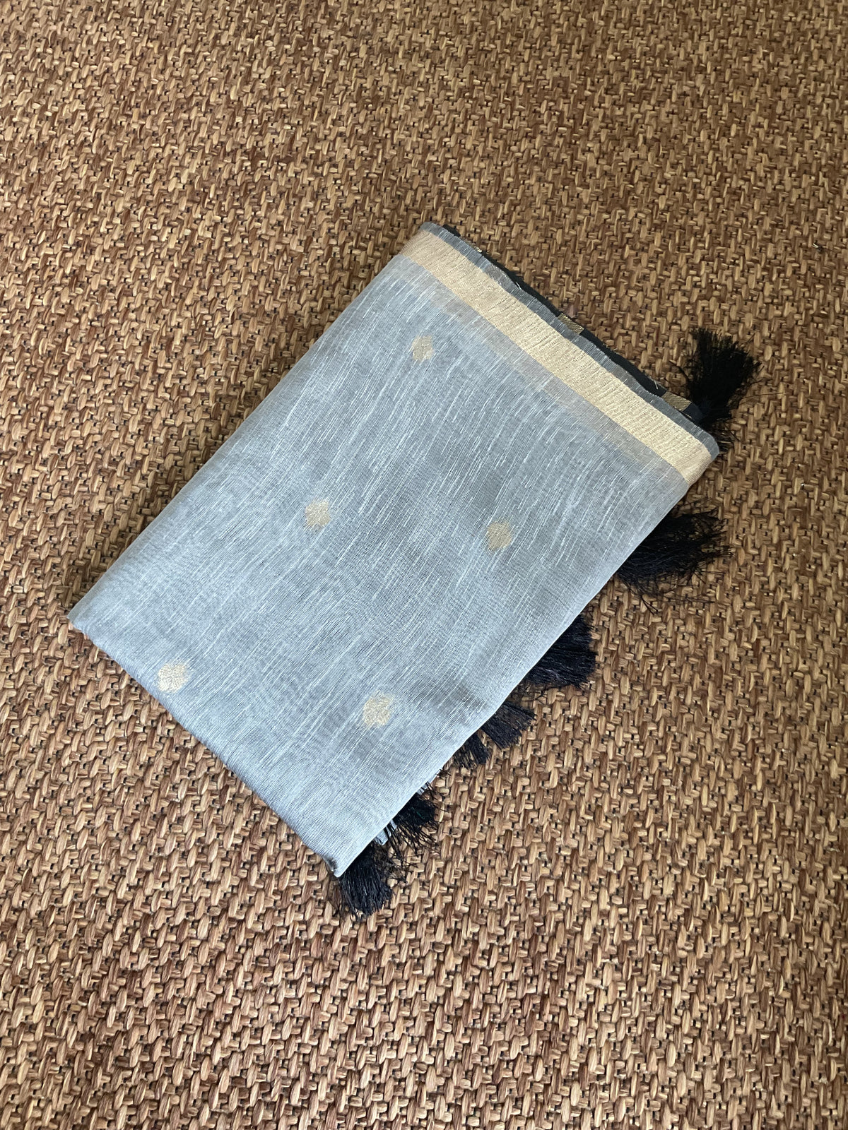 A light blue woven scarf with small gold motifs and black tassels, laid flat on a brown woven rug.