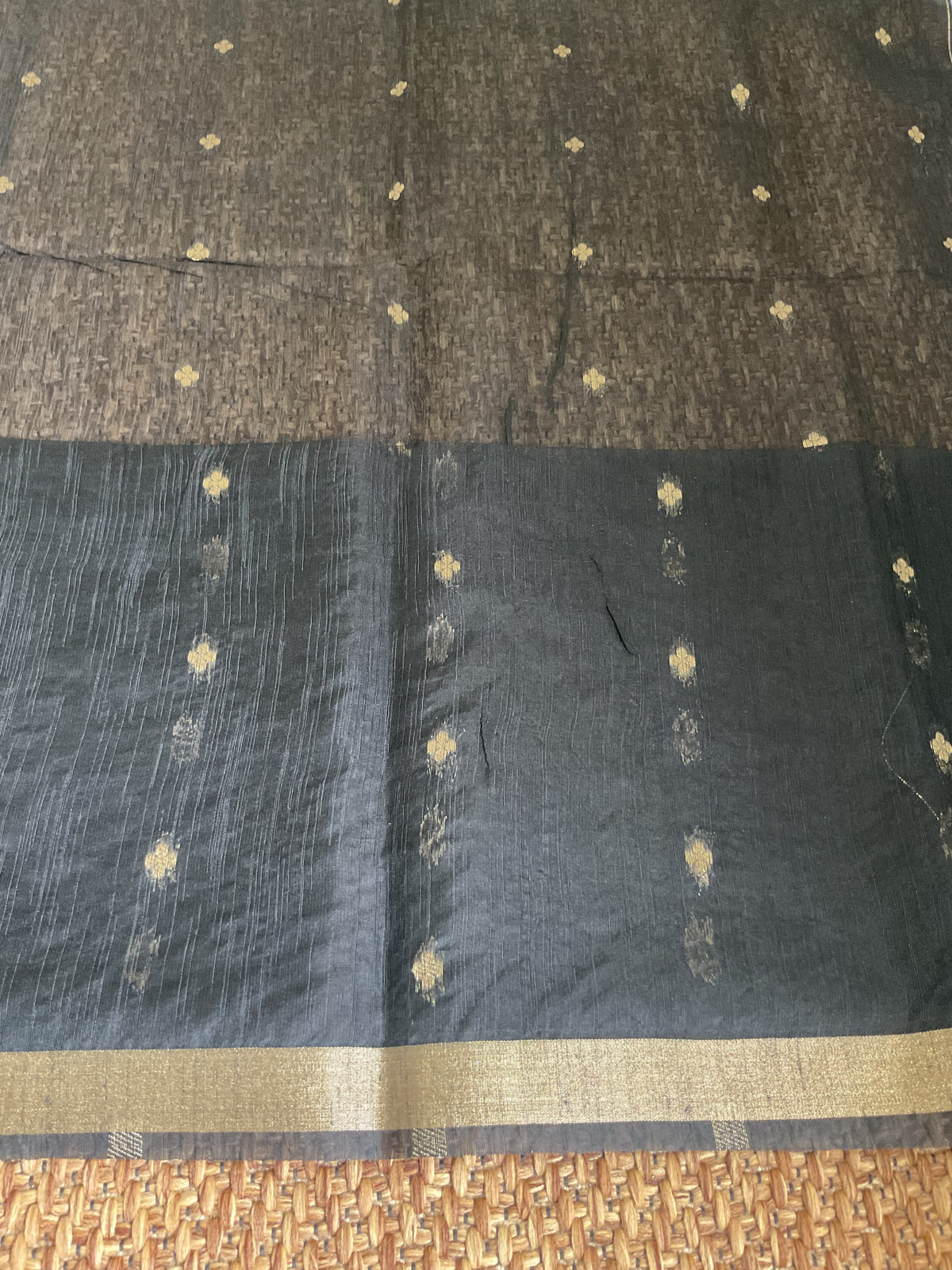 A dark charcoal gray silk saree with a wide gold zari border and small gold motifs, laid flat on a woven mat.